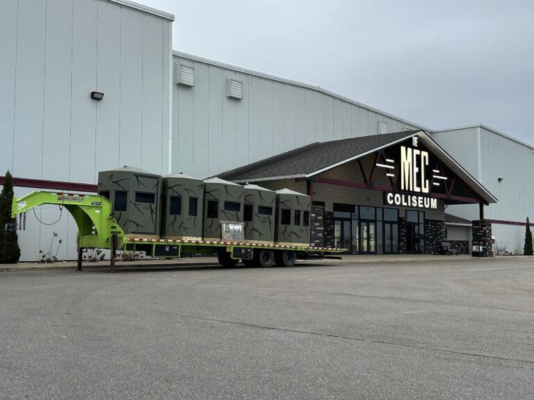 Exterior view of The MEC Coliseum building with hunting blinds on a trailer, marking the location for the Midwest Sportsmen Classic expo in Shipshewana, Indiana.